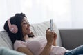 Glad young black lady in wireless headphones typing on phone in living room interior