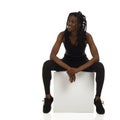 Happy african american woman in black sports clothes is sitting on a white cube. Full length isolated