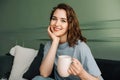 A smiling young woman with a cup of hot drink sits on a couch enjoying some alone time in the living room. Royalty Free Stock Photo