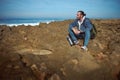 Relaxed bearded male traveler, adventurer, backpacked tourist sitting on a rocky cliff, looking into the distance