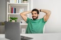 Relaxed, bearded man in a green t-shirt leaning back in his chair with hands behind his head Royalty Free Stock Photo