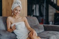 Relaxed beautiful young woman has healthy skin wears soft towel on head and around naked body holds mug of coffee poses at sofa Royalty Free Stock Photo