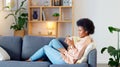 Relaxed Black woman listening to a podcast while drinking coffee and sitting on a couch at home. Happy African American Royalty Free Stock Photo