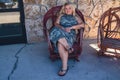 Relaxed blonde woman sits with crossed legs wearing a dress in a rustic rocking chair, outdoors