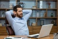 Relaxed brunette man leaning back and holding hands behind head in personal workspace with pc. Calm head of company