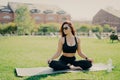 Relaxed brunette woman sits on fitness mat on lotus yoga pose wears sunglasses cropped top and leggings looks aside drinks water