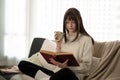 A relaxed caucasian woman is reading a book and sipping coffee on a couch in her living room