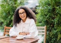Relaxed cheerful Afro woman in white clothes, drinks coffee in outdoor cafeteria, sits at wooden table on chair against green Royalty Free Stock Photo