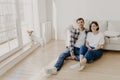 Relaxed couple sit on floor near couch, embrace and smile, dressed in casual clothes and white socks, enjoy domestic atmosphere,