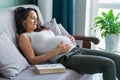 Relaxed dark-haired pregnant woman lying on comfortable grey couch with pillows touching her tummy, smiling and relaxing Royalty Free Stock Photo