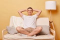 Relaxed delighted brunette adult man wearing casual clothing sitting on sofa against beige wall resting at home keeps hands behind Royalty Free Stock Photo