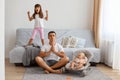 Relaxed father playing with his daughters while sitting on floor near sofa, wearing white t shirt and jeans short, doing yoga Royalty Free Stock Photo