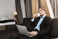 Relaxed handsome businessman with sylish hairstyle and beard sitting in hotel room, drinkig coffee, working on new