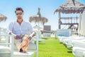 Relaxed handsome man sitting on white chairs during summer at the beach. White shirt and sunglasses. Legs stretched out Royalty Free Stock Photo