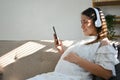 Relaxed Asian pregnant woman listening to music, using her smartphone while resting on sofa Royalty Free Stock Photo