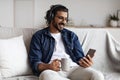 Relaxed Indian Guy Listening Music At Home, Using Smartphone And Wireless Headphones