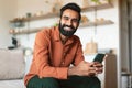 Relaxed Indian Guy Using Smartphone Resting On Couch At Home