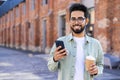 Relaxed middle eastern man holding telephone and plastic coffee cup while walking during daytime. Positive brunet Royalty Free Stock Photo