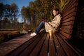 Beautiful peaceful young brunette sitting on wooden bench in park, reading book and drinking coffee, enjoying warm sunny autumn Royalty Free Stock Photo