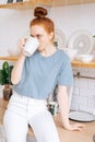 Relaxed redhead young woman holding cup of hot coffee while standing in kitchen room. Royalty Free Stock Photo