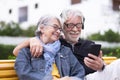 Relaxed senior couple white haired sitting on a yellow bench looking at the same smart phone, bearded man with headphones, Two Royalty Free Stock Photo