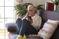 A relaxed senior woman sitting on the sofa at home drinking a coffee cup and looking at her smart phone - attractive woman with Royalty Free Stock Photo