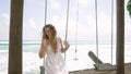 Elegant woman in white dress swings leisurely beside azure sea. Relaxed vacation vibe, breeze plays with wavy hair