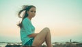 Relaxed woman sitting on stone and posing on shore. Side view of pleasant girl spending time enjoying vacation on stony shore