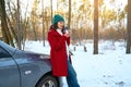 Woman traveler holds thermo mug with hot tea, enjoys coffee break outdoor, sitting on the car hood in snowy woodland. Sun rays