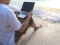 Relaxed young Asian man with laptop sitting on sand of tropical beach. Summer vacations concept. Royalty Free Stock Photo