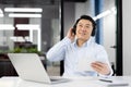 Relaxed young Asian man sitting in the office at the desk wearing headphones, holding a mobile phone, listening to music Royalty Free Stock Photo