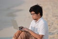 Relaxed young Asian man using laptop on the sandy of beach. Internet of things concept. Royalty Free Stock Photo