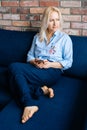 Relaxed young blonde woman using cell phone while sitting on soft dark blue sofa in living room. Royalty Free Stock Photo