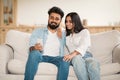 Relaxed young indian couple sitting on sofa at home, using smartphone and smiling, copy space Royalty Free Stock Photo