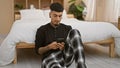 Relaxed young latin man using smartphone while sitting on bedroom floor, comfortably engulfed in cosy morning pyjama vibes Royalty Free Stock Photo