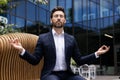 Relaxed young male businessman sitting on bench near office center wearing suit and lotus yoga pose, eyes closed Royalty Free Stock Photo