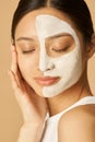 Relaxed young woman with facial mask applied on half of her face receiving spa treatments, posing for camera with eyes