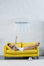 relaxed young woman reading book on couch and pointing at air conditioner hanging on wall Royalty Free Stock Photo