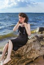 Relaxing Caucasian Brunette Girl Posing in Black Dress On Stone Line At Sea During Sunny Day