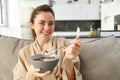 Relaxing morning at home. Young beautiful woman in bathrobe, eating cereals on sofa, having her breakfast in living room Royalty Free Stock Photo
