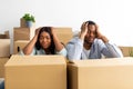 Relocation concept. Frustrated black spouses sitting among cardboard boxes, having problems and packing clothes