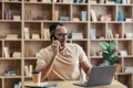 Remote business concept. Concerned arab man talking on cellphone and using laptop while sitting at desk at home Royalty Free Stock Photo