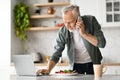 Remote Business. Senior Man Using Cellphone And Working On Laptop In Kitchen Royalty Free Stock Photo