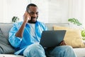 Remote career concept. Happy african american guy talking on cellphone and using laptop, sitting on sofa at home Royalty Free Stock Photo
