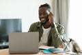 Remote career. Happy african american man talking on cellphone and using laptop, sitting at workplace at home