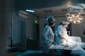 Remote view of male surgeon and African-American female nurse performing operation under bright electric lamp in modern