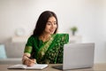 Remote work or education. Lovely Indian woman in sari dress writing down info in notebook in front of laptop at home Royalty Free Stock Photo