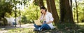 Remote work. Smiling asian girl, student doing homework remotely from park, sitting with laptop near tree and typing Royalty Free Stock Photo