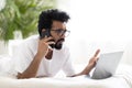 Remote Work Stress. Young indian man using cellphone and laptop in bed Royalty Free Stock Photo