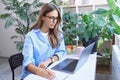 Remote work. Young attractive business woman in glasses sitting at desk, using laptop and making some notes, she is Royalty Free Stock Photo